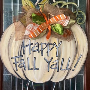 Pode incluir: Um pendurador de porta de ab&oacute;bora branca com um la&ccedil;o de juta marrom e uma fita xadrez verde e laranja. A ab&oacute;bora tem as palavras "Happy Fall Y'all!" pintadas nela.