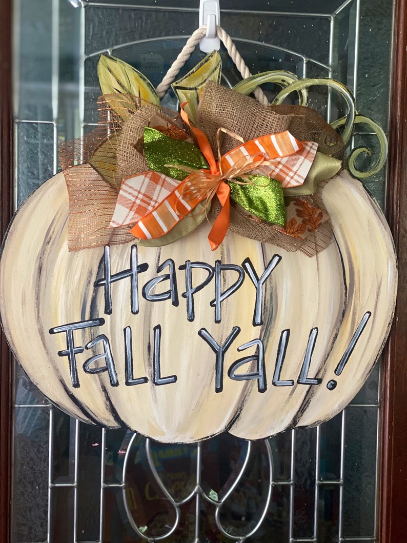 Pode incluir: Um pendurador de porta de ab&oacute;bora branca com um la&ccedil;o de juta marrom e uma fita xadrez verde e laranja. A ab&oacute;bora tem as palavras "Happy Fall Y'all!" pintadas nela.