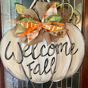 Pode incluir: Um pendurador de porta em forma de ab&oacute;bora branca com as palavras "Welcome Fall" pintadas em preto e verde. A ab&oacute;bora tem um grande la&ccedil;o de juta com fitas laranja, verde e marrom.
