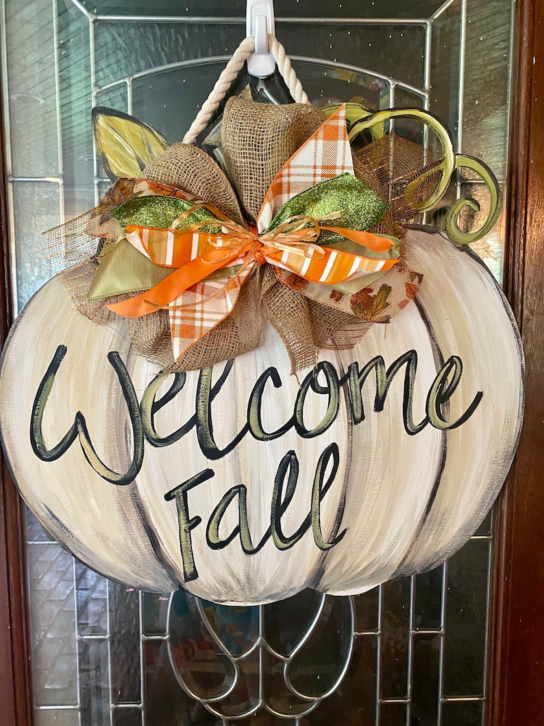 Pode incluir: Um pendurador de porta em forma de ab&oacute;bora branca com as palavras "Welcome Fall" pintadas em preto e verde. A ab&oacute;bora tem um grande la&ccedil;o de juta com fitas laranja, verde e marrom.