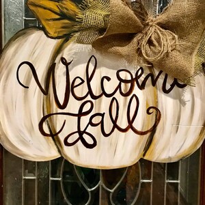 Pode incluir: Um pendurador de porta em forma de ab&oacute;bora branca com as palavras "Welcome Fall" pintadas em marrom. Um la&ccedil;o de juta est&aacute; amarrado no topo da ab&oacute;bora.