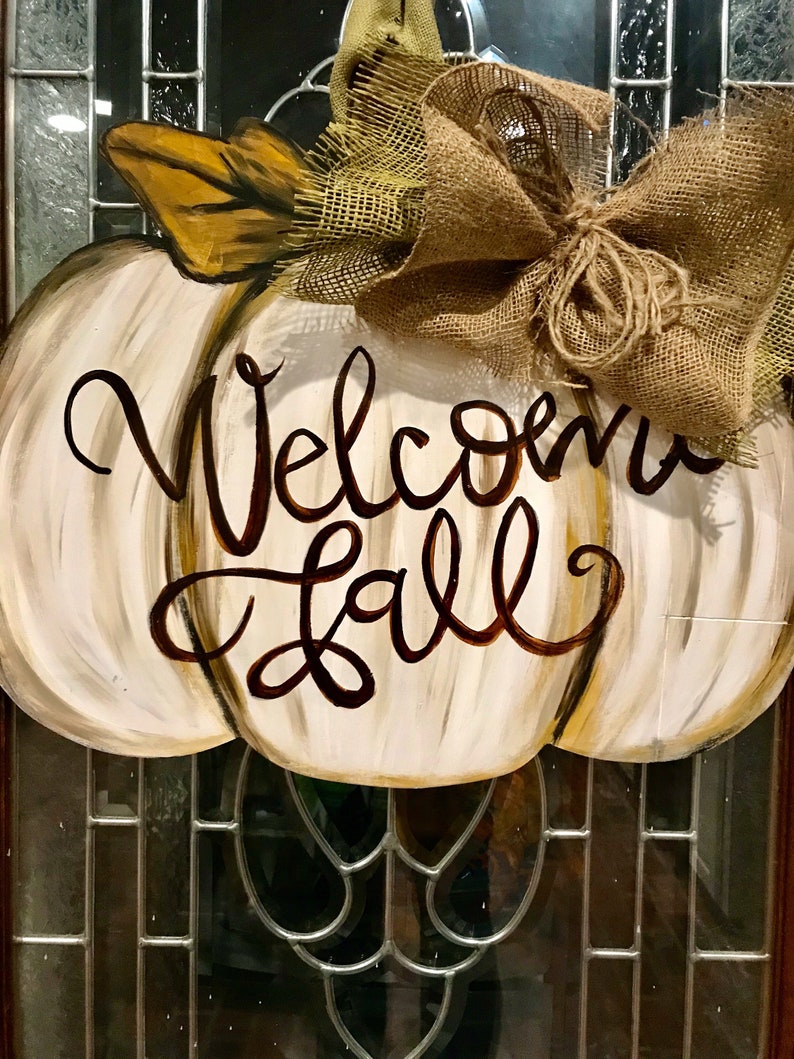 Pode incluir: Um pendurador de porta em forma de ab&oacute;bora branca com as palavras "Welcome Fall" pintadas em marrom. Um la&ccedil;o de juta est&aacute; amarrado no topo da ab&oacute;bora.