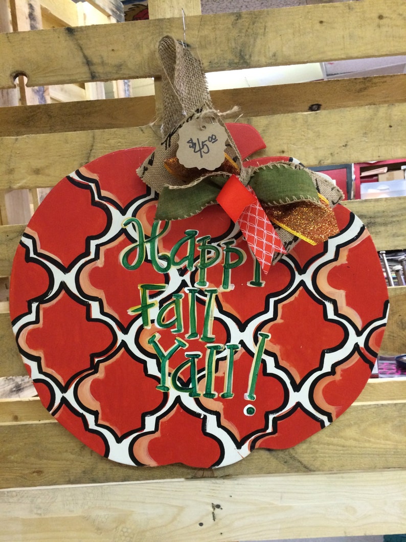 Pode incluir: Um pendurador de porta em forma de ab&oacute;bora vermelho e branco com um padr&atilde;o geom&eacute;trico. As palavras "Happy Fall Y'all!" est&atilde;o escritas em verde na frente. Um la&ccedil;o de juta com uma etiqueta de pre&ccedil;o que diz "45 &euro;" est&aacute; preso ao topo.