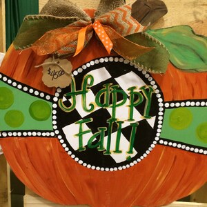 Pode incluir: Um pendurador de porta de ab&oacute;bora pintado com um fundo xadrez preto e branco e as palavras "Happy Fall" em letras douradas. A ab&oacute;bora &eacute; decorada com uma fita verde e branca e um la&ccedil;o de juta.