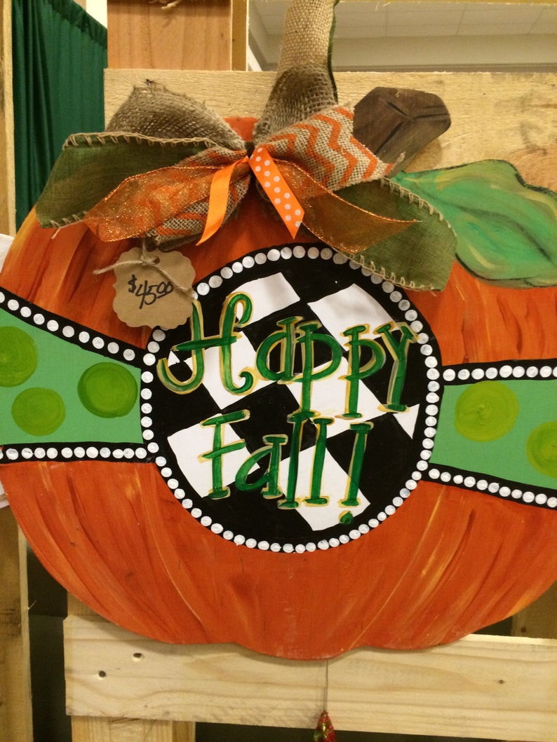 Pode incluir: Um pendurador de porta de ab&oacute;bora pintado com um fundo xadrez preto e branco e as palavras "Happy Fall" em letras douradas. A ab&oacute;bora &eacute; decorada com uma fita verde e branca e um la&ccedil;o de juta.