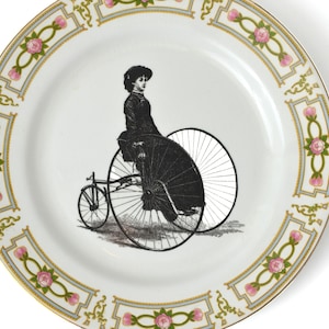 May include: White ceramic plate featuring a black and white illustration of a woman riding a high-wheel bicycle. The plate's rim is decorated with pink roses and gold trim, creating a vintage aesthetic. A decorative piece for display.