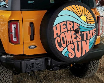 May include: A yellow Ford Bronco with a spare tire cover that says "Here Comes The Sun" in a retro font with a sun and wave design.