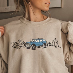 May include: A light beige sweatshirt with a blue graphic of a jeep driving through a mountain range.