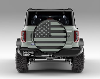 May include: A gray Ford Bronco with a spare tire cover featuring a gray and black American flag design. The spare tire cover has the text "MUD DIGGER DESIGN CO." printed on it.