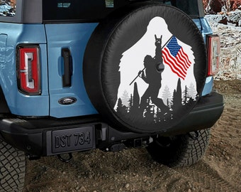 May include: A blue Ford Bronco with a spare tire cover featuring a silhouette of Bigfoot holding an American flag. The spare tire cover is black with white accents. The license plate reads "DST 734".