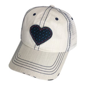 May include: A cream-colored baseball cap featuring a dark blue heart-shaped patch with a light blue star pattern. The cap has dark blue stitching, a distressed brim, and a mesh back panel. The design includes dark blue eyelets on the crown.