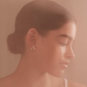 May include: A woman with dark hair wearing a pair of small silver triangle earrings.