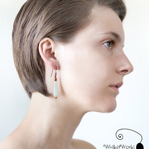 May include: A woman with short, straight brown hair is wearing a pair of dangle earrings with a light green stone and copper beads. The earrings are hanging from her left ear.