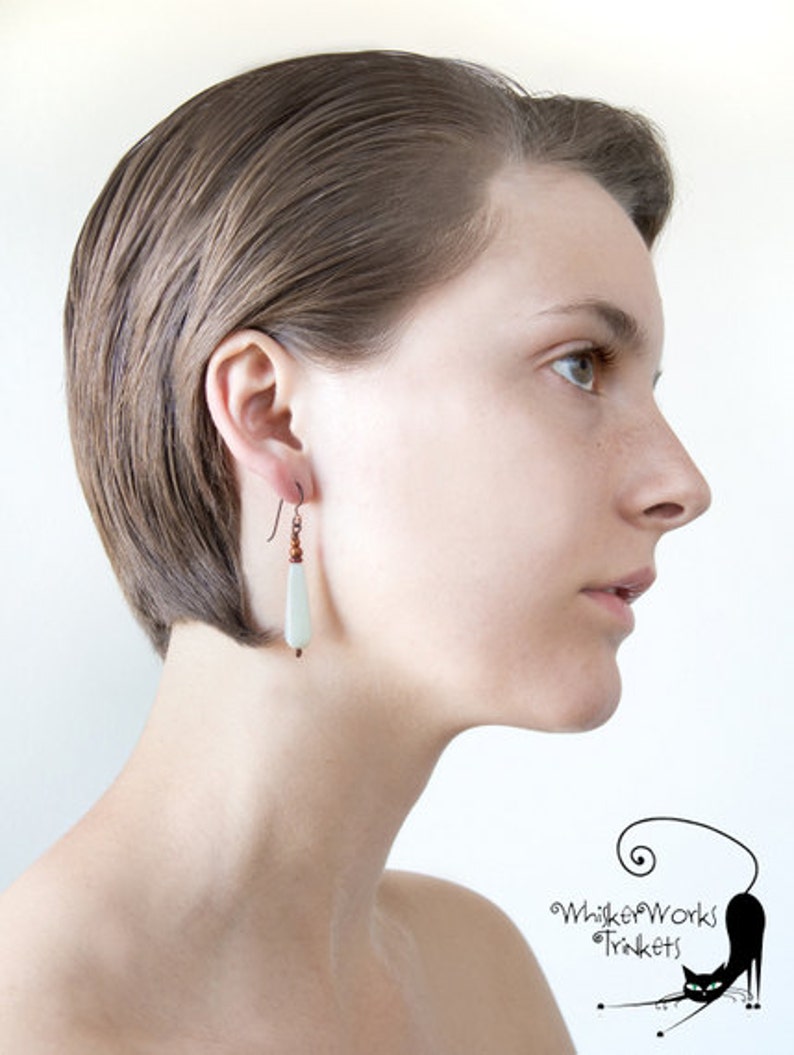 May include: A woman with short, straight brown hair is wearing a pair of dangle earrings with a light green stone and copper beads. The earrings are hanging from her left ear.
