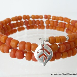 Könnte beinhalten: Ein dreifach gewickeltes Armband aus orangefarbenen Glasperlen mit einem silbernen Blattanhänger.