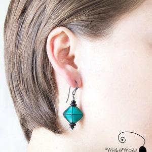 May include: Teal and black geometric dangle earrings with a black wire hook. The earrings are worn by a person with short brown hair.