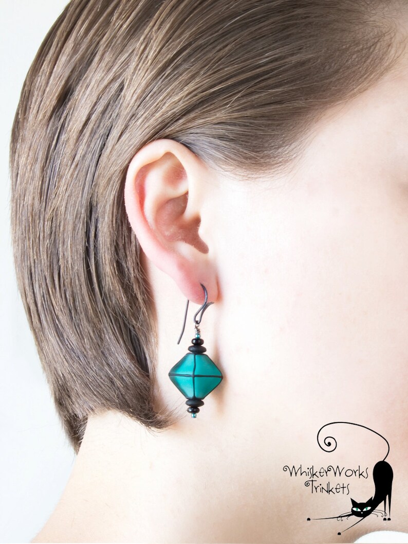 May include: Teal and black geometric dangle earrings with a black wire hook. The earrings are worn by a person with short brown hair.