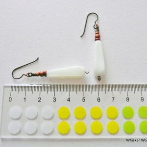 May include: A pair of white teardrop-shaped earrings with copper beads and wire. The earrings are shown next to a ruler with markings in inches.