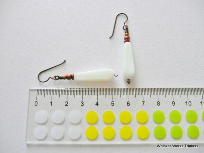 May include: A pair of white teardrop-shaped earrings with copper beads and wire. The earrings are shown next to a ruler with markings in inches.