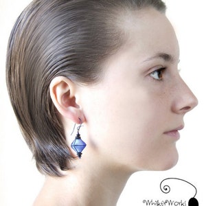 May include: A woman with short, straight, dark hair is wearing a pair of blue, diamond-shaped earrings. The earrings are made of glass and have a silver wire.