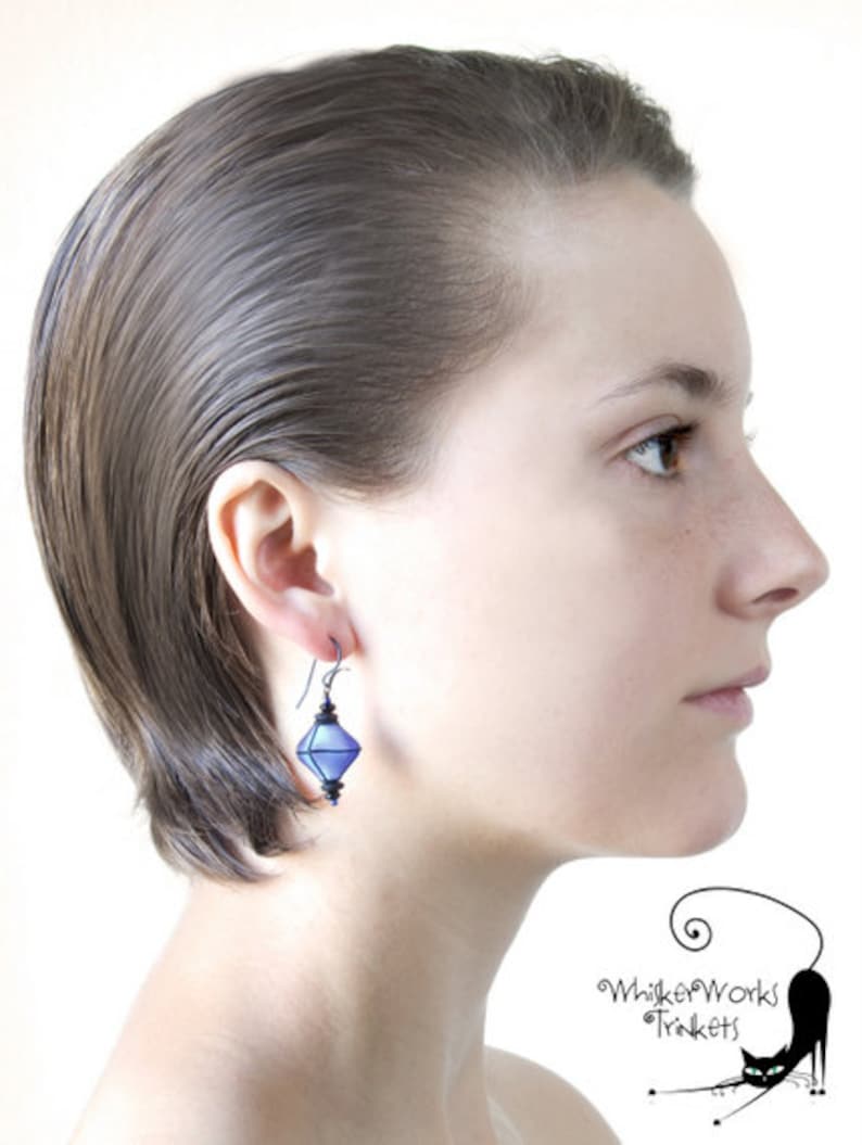 May include: A woman with short, straight, dark hair is wearing a pair of blue, diamond-shaped earrings. The earrings are made of glass and have a silver wire.