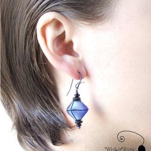 May include: A pair of dangle earrings with a geometric, faceted, blue glass pendant. The pendant is accented with black and blue beads. The earrings are made with a silver-toned hook.