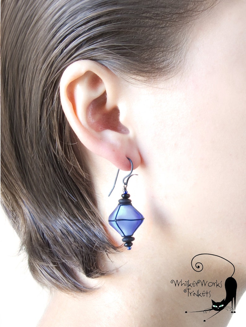 May include: A pair of dangle earrings with a geometric, faceted, blue glass pendant. The pendant is accented with black and blue beads. The earrings are made with a silver-toned hook.