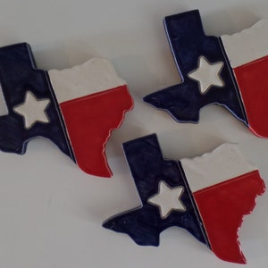 May include: Three ceramic Texas-shaped coasters with the Texas flag design. The coasters are blue, white, and red with a white star in the center.