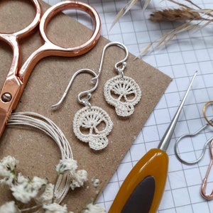 May include: A pair of white crocheted skull earrings with silver hooks. The earrings are displayed on a brown surface with a grid pattern.