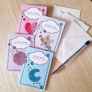 May include: Four colourful cards with crochet pattern stickers. The cards are pink, blue, purple, and light blue. Each card features a different crochet pattern sticker: a heart, a flower, a star, and a moon. The cards have the text "Little Heart", "Little Flower", "Little Star", and "Little Moon" on them.