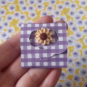May include: A small purple and white gingham paper envelope with a crocheted sunflower in the center. The sunflower is yellow with a brown center.