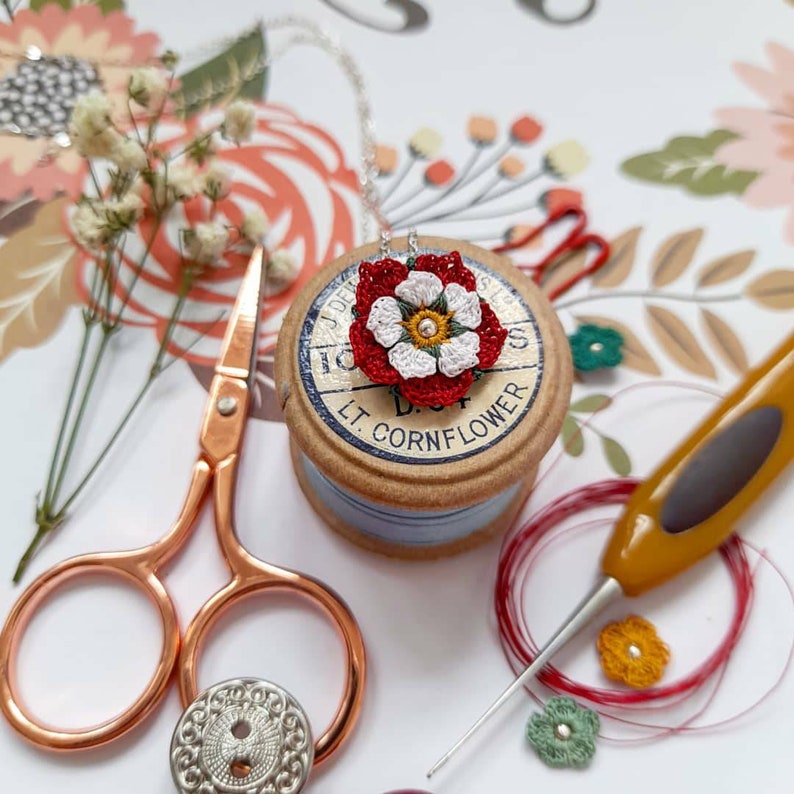 Op de afbeelding: Een close-up van een witte en rode gecrochete bloemhanger aan een zilveren ketting. De hanger ligt op een spoel garen met het label "D.L.T. Cornflower".