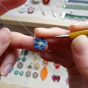 K&ouml;nnte beinhalten: Nahaufnahme von jemandes H&auml;nden, die eine kleine blaue Blume mit gelber Mitte h&auml;keln. Die Blume wird mit einer Nadel und Garn gehalten.