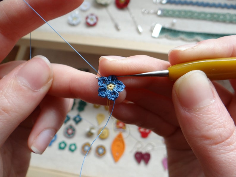 K&ouml;nnte beinhalten: Nahaufnahme von jemandes H&auml;nden, die eine kleine blaue Blume mit gelber Mitte h&auml;keln. Die Blume wird mit einer Nadel und Garn gehalten.