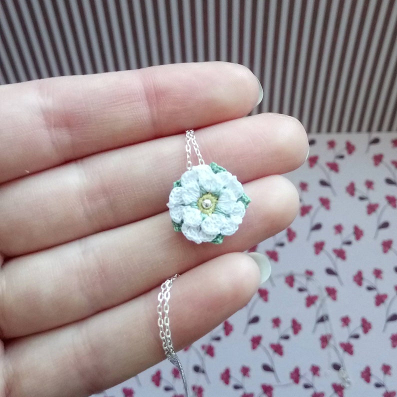 Puede incluir: Un delicado collar con un colgante de flor blanca hecha a crochet y una cadena de plata. La flor tiene un centro verde con una peque&ntilde;a perla plateada.