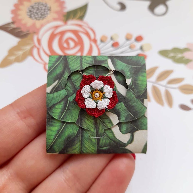 Op de afbeelding: Een witte en rode gecrochete bloemhangerketting met een zilveren ketting. De hanger bevindt zich op een kaart met een groen en wit bloemenpatroon.