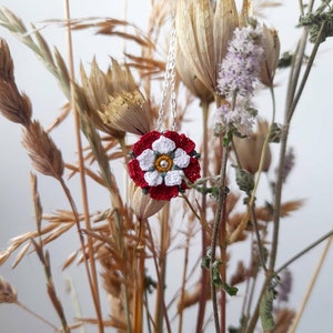 Op de afbeelding: Een witte gehaakte bloem met een rood centrum en groene accenten hangt aan een zilveren ketting. De bloem wordt omringd door gedroogde grassen en bloemen.
