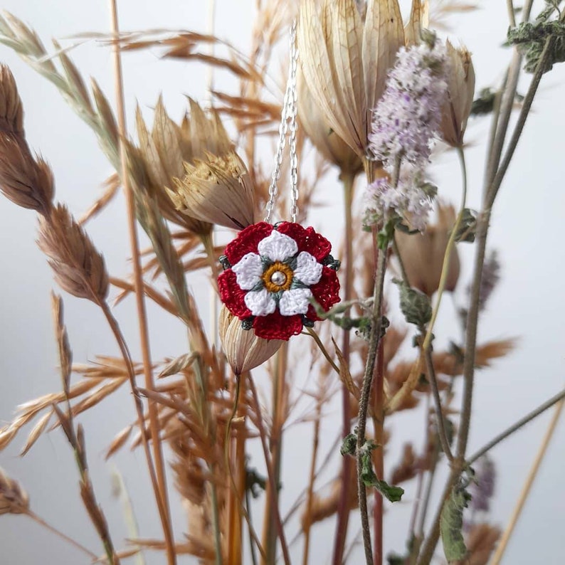 Op de afbeelding: Een witte gehaakte bloem met een rood centrum en groene accenten hangt aan een zilveren ketting. De bloem wordt omringd door gedroogde grassen en bloemen.