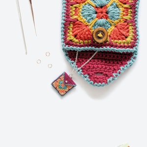 K&ouml;nnte beinhalten: Eine geh&auml;kelte Tasche mit einem farbenfrohen geometrischen Muster und einem Knopfverschluss. Eine kleine geh&auml;kelte Tasche mit einem &auml;hnlichen Muster h&auml;ngt an einer silbernen Kette.
