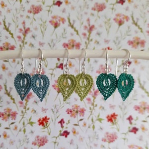 May include: Three pairs of crocheted leaf earrings hanging on a white stand. The earrings are in shades of teal, green, and dark green.