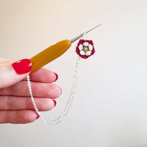 Op de afbeelding: Een gehaakte witte bloem met een rood hart en groene bladeren is bevestigd aan een zilveren ketting. Een gele haaknaald wordt gebruikt om de ketting vast te houden.