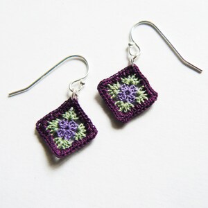 May include: A pair of purple crochet earrings with green and purple flower accents. The earrings are attached to silver hooks.