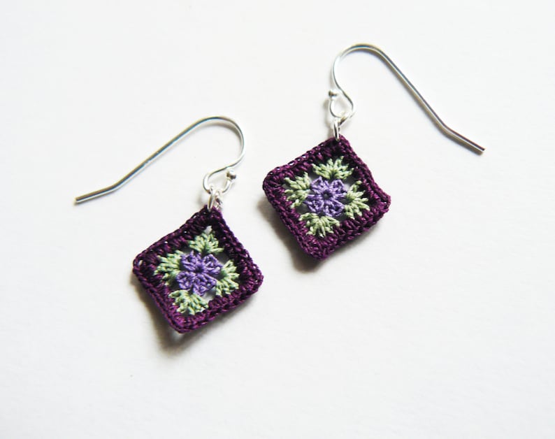 May include: A pair of purple crochet earrings with green and purple flower accents. The earrings are attached to silver hooks.