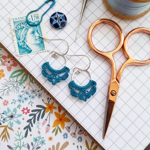 May include: A pair of silver hoop earrings with teal crocheted embellishments. The earrings are on a white gridded surface.