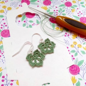 May include: A pair of green crochet flower earrings with silver hooks. The earrings are on a white background with a floral pattern.
