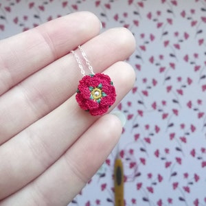 Puede incluir: Un delicado collar de cadena plateada con un colgante de flor roja de ganchillo. La flor tiene hojas verdes y un centro de perla blanca.