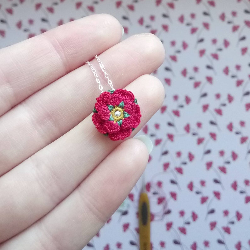 Puede incluir: Un delicado collar de cadena plateada con un colgante de flor roja de ganchillo. La flor tiene hojas verdes y un centro de perla blanca.