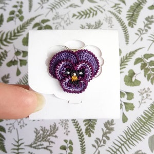 May include: A purple and white crocheted pansy flower pendant on a silver chain. The flower has a yellow center and black eyes.