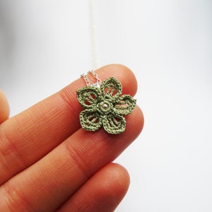 May include: A handmade, olive green crochet flower pendant necklace. The flower has five petals and a small, silver-colored bead in the center. The necklace chain is a delicate silver color. The flower is approximately 1.5 inches in diameter.