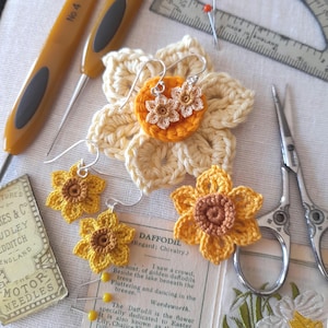 May include: Crocheted daffodil earrings with yellow and brown petals. The earrings are displayed on a crocheted daffodil flower and a vintage needle packaging.